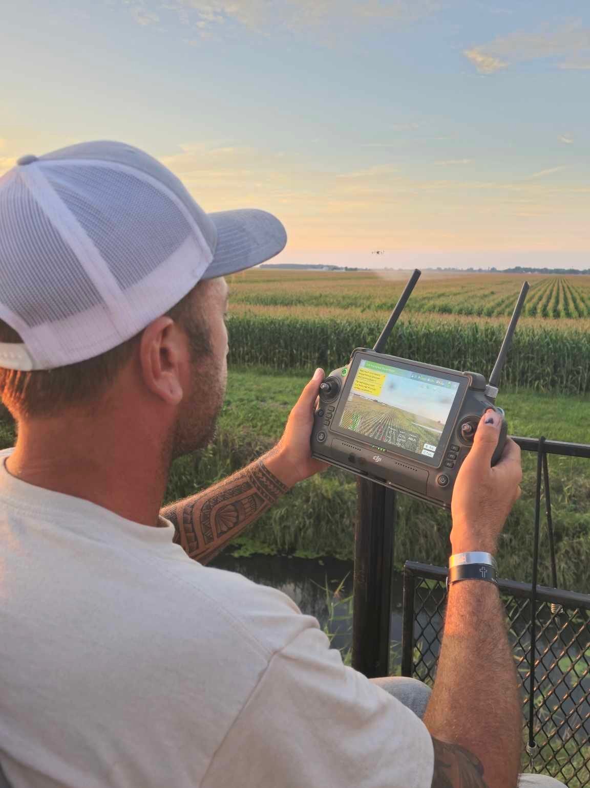 Heritage Drone Service drone pilot operating an agricultural drone controller during a crop spraying mission at sunset.
